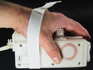 Palm Writer Being Gripped In Side Perspective View Showing Wrist Strap, Wrist Rest, Suspension Strap, Tactile Feedback Unit or Writer, and Charge Coil on Left Plate